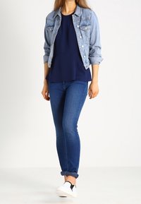 Frau in einer hellen Jeansjacke, dunkelblauem Blusenshirt, dunkelblauen Jeans, die an den Säumen aufgerollt sind, und weißen Slippern vor einem weißen Hintergrund.