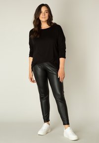 Zwarte langemouwen top gecombineerd met nauwsluitende zwarte neplederen leggings. Model draagt witte sneakers en staat op een neutrale achtergrond.