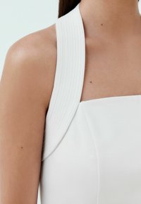 Vit klänning med en bred, ribbad halterneck. Slät textil med strukturerad axeldesign, närbild som visar axel- och nackområde.