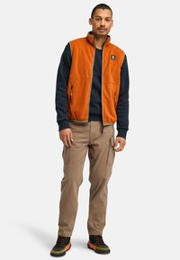 Orange fleece vest with high collar, navy sweatshirt underneath, beige cargo pants, and black shoes with red and green accents.