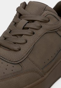 Καφέ δερμάτινο αθλητικό παπούτσι με υφή στο πάνω μέρος, επενδυμένο κολάρο, στρογγυλή μύτη και επίπεδα κορδόνια. Διαθέτει διάτρητη πλευρική επιφάνεια και ραφές με αντίθεση.