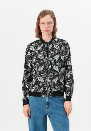 Vrouw in een zwarte bloemen bomberjack met rits en ribgebreide manchetten en kraag, gecombineerd met lichtblauwe jeans, staande tegen een witte achtergrond.