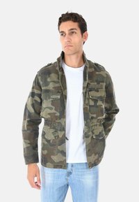 Giacca camouflage con chiusura con zip, diverse tasche frontali, epaulettes sulle spalle con bottoni, nei toni di verde, marrone e beige. Indossata sopra una maglietta bianca.
