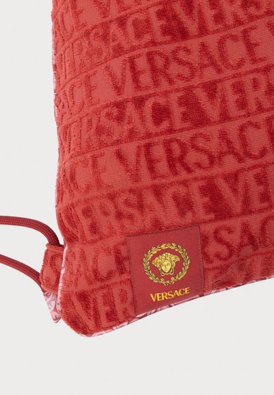 Sarkana dekoratīvā spilvena ar reljefu rakstu, kurā redzams uzraksts "VERSACE", teksturētu virsmu un logotipa uzlīmi ar Medūzas dizainu.