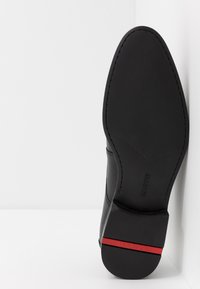 Semelle de chaussure en cuir noir avec texture lisse et bout pointu ; présente un accent rouge près du talon et un design perforé sur les côtés.