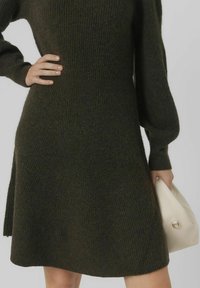 Robe en maille vert foncé avec texture côtelée, cintrée à la taille et évasée vers l'ourlet. Associée à un sac à main de couleur crème.