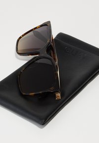 Tortoiseshell sunglasses with dark lenses, featuring a geometric shape and gold hardware, positioned on a black leather case embossed with "QUAY."