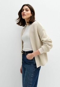 Ljus beige kardiagan med texturerat mönster, öppen framsida och sänkta axlar, bärs över en åtsittande vit topp och blå högmidjade jeans.