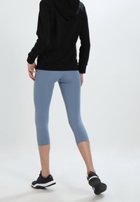 Sweat-shirt à capuche noir avec une coupe ajustée, associé à des leggings bleu clair trois-quarts. Le mannequin porte des baskets noires. Tissu lisse et extensible.