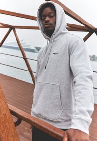 Grå huvtröja med framficka, snodd och logotyp på bröstet. Slät textur, använd i en utomhusmiljö nära vatten.