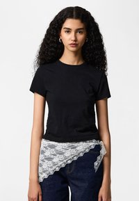 Donna con capelli lunghi e ricci che indossa una maglietta nera a maniche corte e jeans scuri con un sovrapposto asimmetrico in pizzo bianco intorno alla vita.