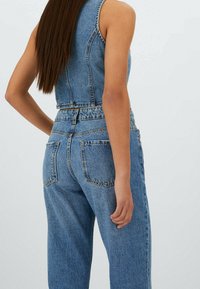 Kvinna med långt brunt hår som bär en ärmlös blå denimväst och höga blå denimjeans, står mot en enkel bakgrund.