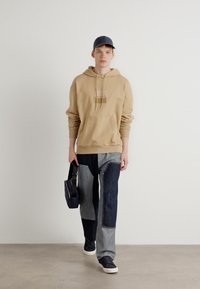 Felpa con cappuccio marrone e logo, jeans scuri e pantaloni in denim chiaro con toppe, cappellino blu, con una borsa nera e scarpe da ginnastica scure.