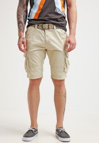 Shorts cargo beiges avec plusieurs poches latérales, fabriqués en coton. Portés avec un t-shirt graphique et des chaussures grises à enfiler.