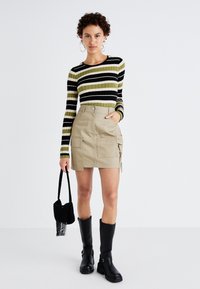 Csíkos, fekete, zöld és fehér hosszú ujjú felső; bézs farmer miniszoknya zsebekkel; fekete, térd fölé érő csizmák; fekete táska.