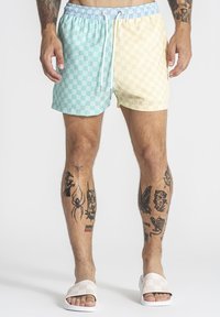Mann trägt pastellblaue und gelbe karierten Badeshorts mit weißem Kordelzug, weiße karierte Slides und sichtbare Bein-Tattoos.