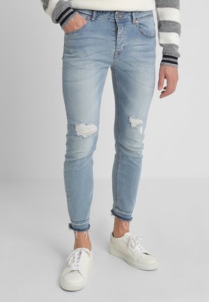 Jean skinny bleu clair déchiré avec ourlets effilochés, porté avec des baskets blanches et un pull rayé gris et blanc.