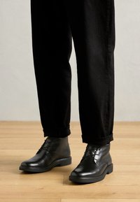 Black leather ankle boots with a smooth finish, closed lacing, and rounded toe, paired with rolled black denim pants on a wooden floor.
