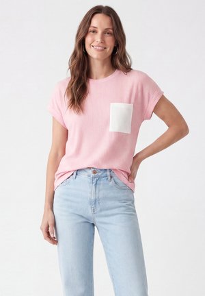 Kvinde smilende iført en lyserød kortærmet ribbet skjorte med en hvid lomme og lyseblå højtaljede jeans mod en ensfarvet baggrund.