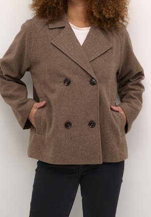 Vrouw draagt een bruine wollen jas met dubbele rij zwarte knopen, handen in de zakken, gecombineerd met een zwarte broek en een wit overhemd eronder.