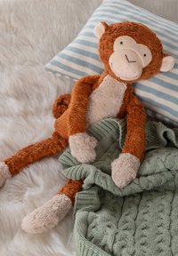 Plush monkey toy with brown fur, beige accents, and long limbs, resting on a striped blue pillow with a green knitted blanket nearby.