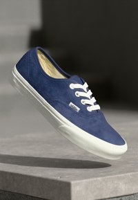 Blue suede low-top sneaker with white rubber sole and laces, featuring a small logo tag on the side. Smooth leather-like texture.