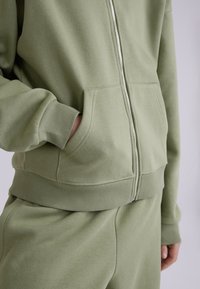 Felpa con zip verde oliva realizzata in tessuto morbido, con polsini e orlo a costine, tasca anteriore e finitura liscia.