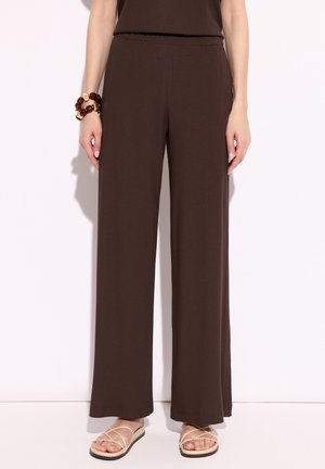 Kobieta w brązowych spodniach o szerokich nogawkach, beżowych sandałach na paski i masywnych brązowych i złotych bransoletkach na lewym nadgarstku.