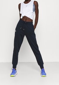 Marina joggerbyxor med elastisk midja och dragsko, kombinerade med en vit kortärmad tanktopp med en knutdetalj. Blå och lila sneakers.