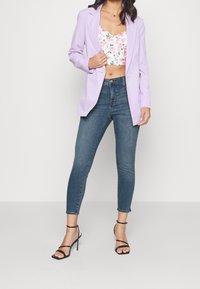 Lavenderkavaj över en blommig croptop, kombinerad med blåa skinny jeans och svarta remklackar. Mjukt tyg och figurnära design genomgående.