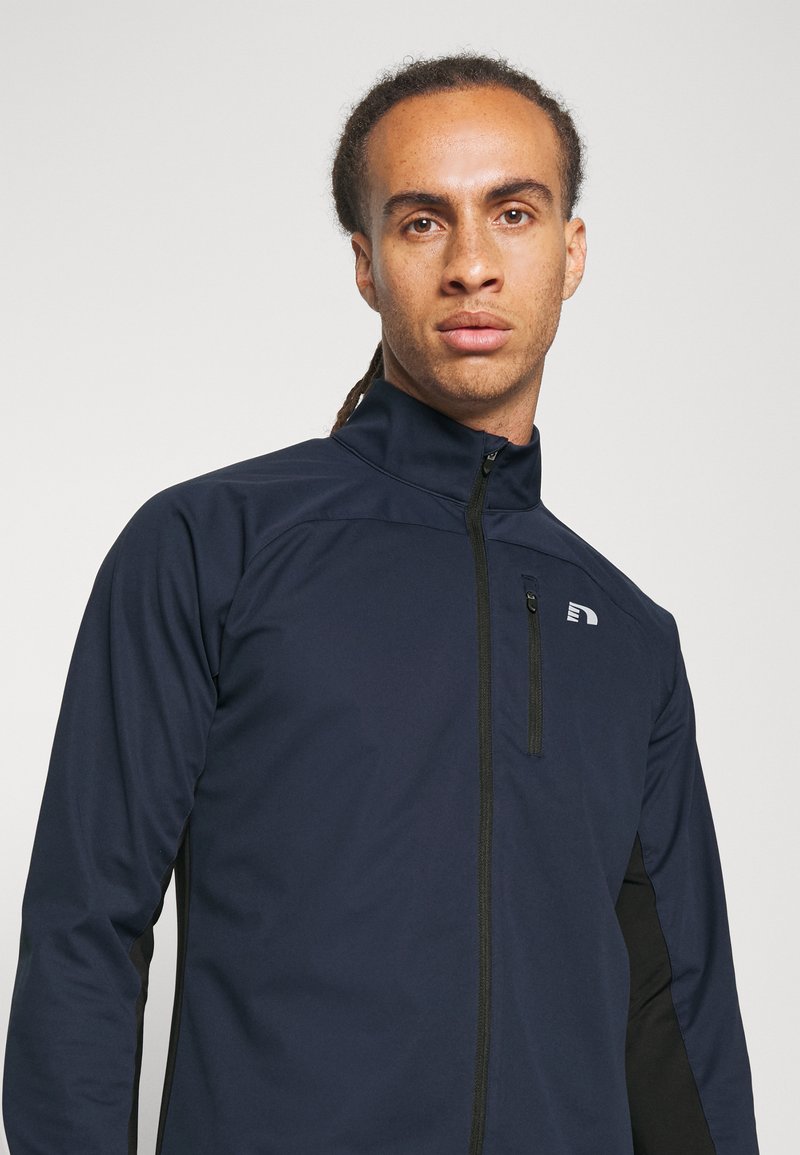 Marineblaues Zip-Jackett mit hohem Kragen, mit schwarzen Seiteneinsätzen und kleinem Logo auf der linken Brust. Hergestellt aus glattem, dehnbarem Stoff.