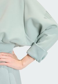 Light blue-green oversized sweatshirt with gathered sleeves and ribbed waistband. Features logo detail on the sleeve. Soft textile finish.