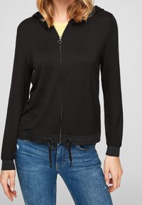 Femme portant un sweat à capuche noir zippé avec des cordons à la taille et un jean bleu, debout devant un fond clair uni.