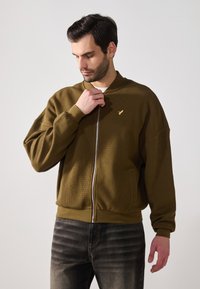 Oliven grønn zip-up jakke med teksturert stoff, ribbede mansjetter og nederkant. Har en liten gulllogo på brystet. Brukt med mørke jeans.