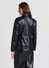 Veste en similicuir noir à la coupe ajustée, ornée de clous argentés sur les manches, avec une silhouette élégante et un col pointu.