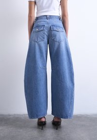 Γυναίκα που φοράει μπλε τζιν παντελόνι με φαρδιά μπατζάκια, λευκό εφαρμοστό τοπ και μαύρες ψηλοτάκουνες σανδάλες, στέκεται σε γκρι δάπεδο μπροστά σε λευκό τοίχο.