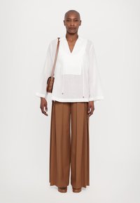 Blusa leggera bianca con scollo a V e maniche lunghe, abbinata a pantaloni marroni a gamba larga. Borsa a spalla marrone con finitura testurizzata.