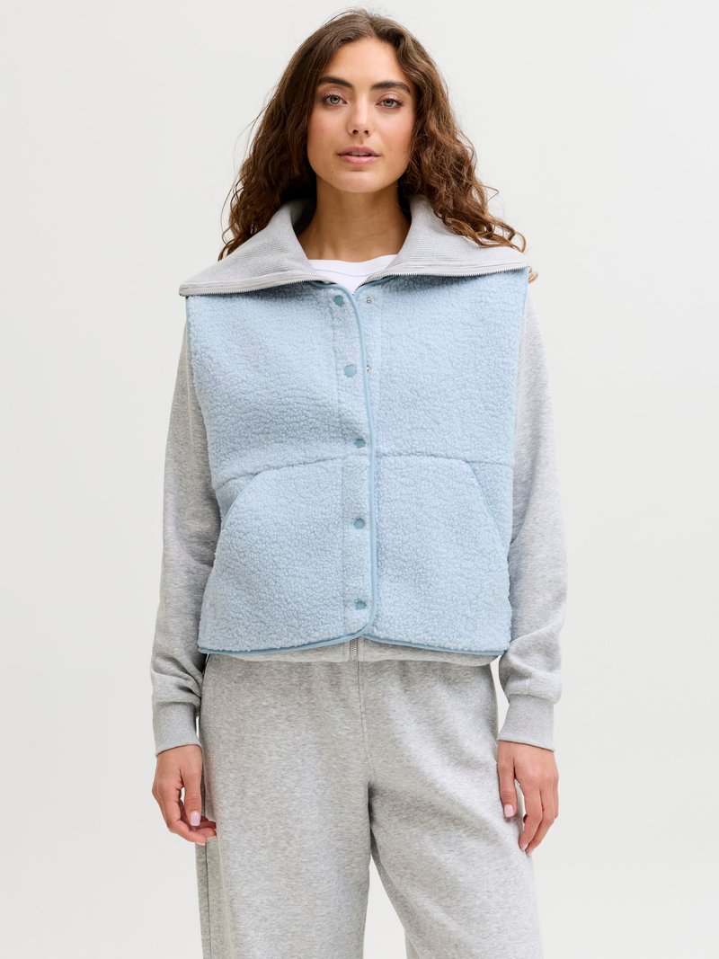 Νεαρή γυναίκα που φοράει ένα γαλάζιο φλις γιλέκο πάνω από γκρι φούτερ και ταιριαστά γκρι φόρμες, στέκεται μπροστά σε απλό λευκό φόντο.