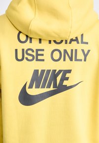 Sárga kapucnis pulóver elülső zsebbel, erőteljes fekete felirattal: "CSAK HIVATALOS HASZNÁLATRA" és a Nike logó jól láthatóan.