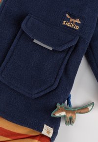 Marineblaue Fleecejacke mit einer aufgesetzten Tasche, die einen Fuchs-Anhänger besitzt. Enthält ein hellbraunes Etikett mit einem Logo und einem rot-orangenen gestreiften Stoff darunter.