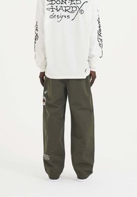 Wit overhemd met lange mouwen en zwarte letters op de achterkant, gecombineerd met ruime olijfgroene broek met een geborduurde patch en witte afwerking.