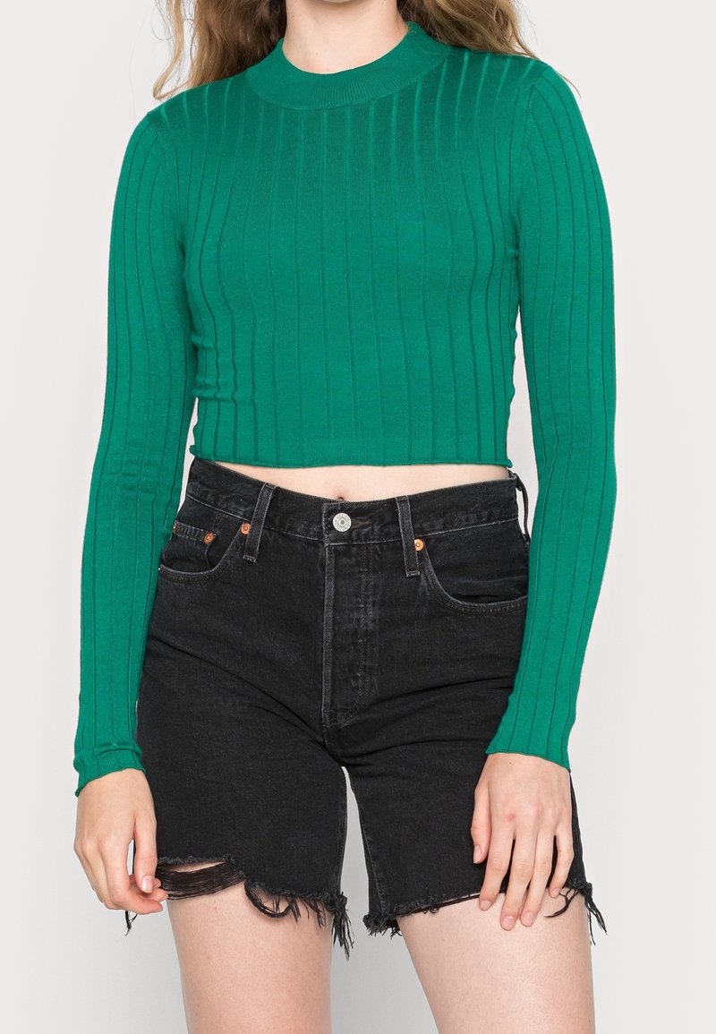 Femme portant un crop top vert côtelé à manches longues et un short en jean noir taille haute avec des ourlets effilochés.