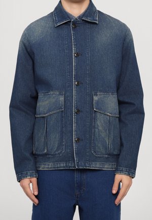 Mies, jolla on sininen denimtakin napitettu versio, jossa on kaksi suurta etutaskua läpillä ja haalistuneet yksityiskohdat, yhdistettynä sinisiin farkkuihin.