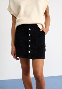 Jupe en velours côtelé noire avec une taille haute, des poches avant et des boutons-pression en métal. Associée à un haut en tricot sans manches de couleur crème.