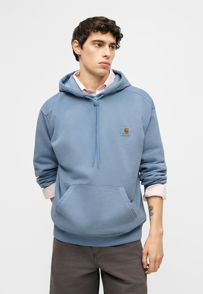 Junger Mann mit lockigem Haar, der einen blauen Carhartt-Kapuzenpullover über einem rosa Hemd trägt und mit den Händen hinter dem Rücken dasteht.