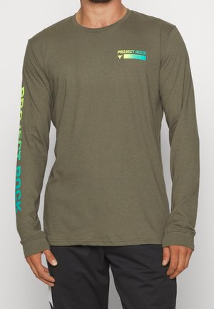 Mann trägt ein olivgrünes Langarmshirt mit dem Schriftzug "Project Rock" auf der linken Brust und am rechten Ärmel, kombiniert mit einer schwarzen Hose.