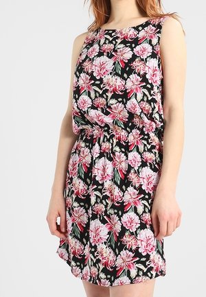 Mujer con vestido negro sin mangas con estampado floral rosa y blanco y cintura fruncida, de pie frente a un fondo liso.