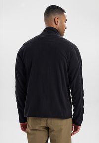Giacca in pile nera con colletto alto, maniche lunghe e una texture morbida. Presenta una zip all'orlo e una vestibilità comoda.