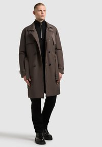 Brown trench coat with double-breasted design, black buttons, and shoulder epaulettes, worn over a black zip-neck knit top and black pants.