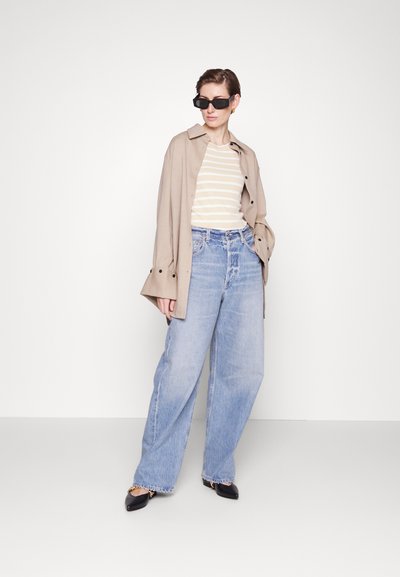 Manteau trench beige, t-shirt rayé blanc et beige, jean large bleu clair, et lunettes de soleil noires avec des ballerines pointues, se tenant devant un fond uni.